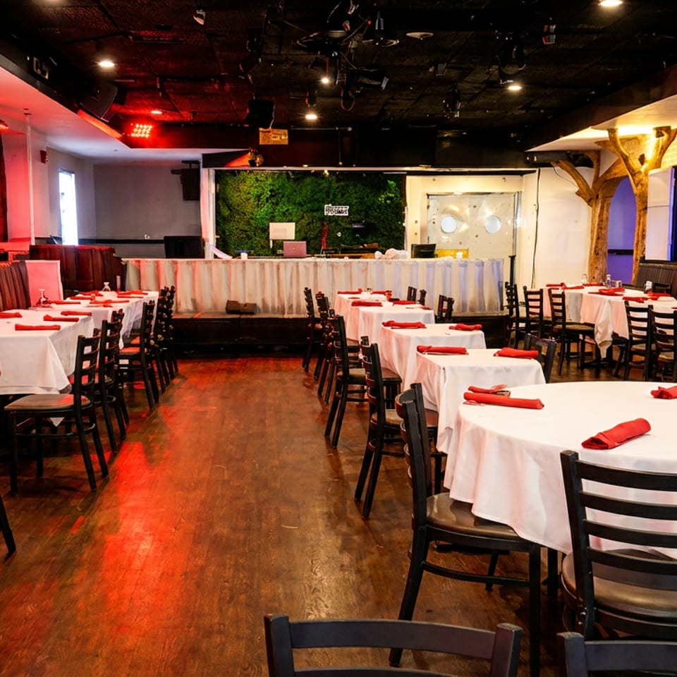 Spacious restaurant dining room with set tables, booths, and a stage, illuminated by red lights.