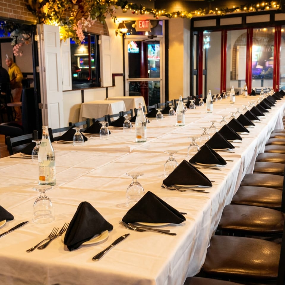 Long banquet table set with white tablecloths, black napkins, and glasses in a warm restaurant.
