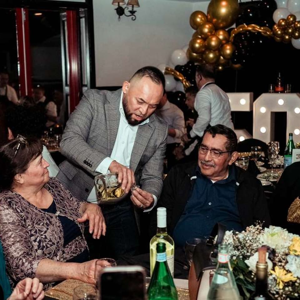 Man in a suit pours a drink for an older man at a lively restaurant celebration.