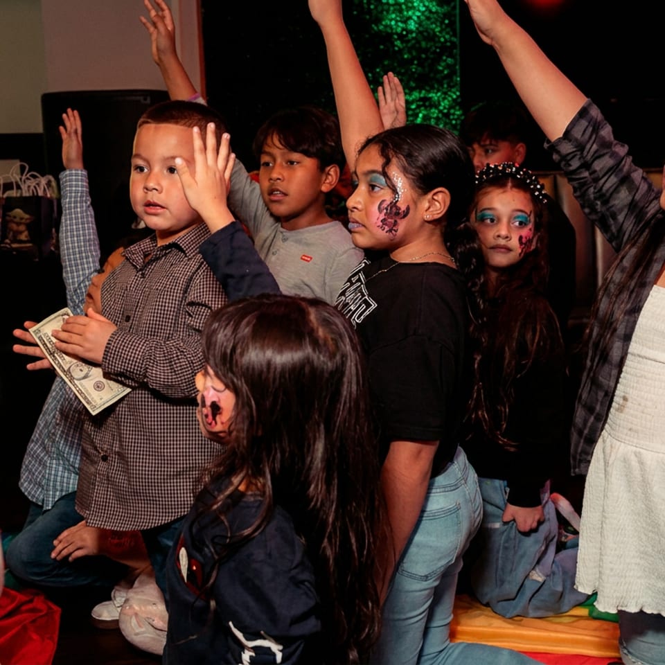 Kids with face paint raise hands at a party, one boy holding a dollar bill.